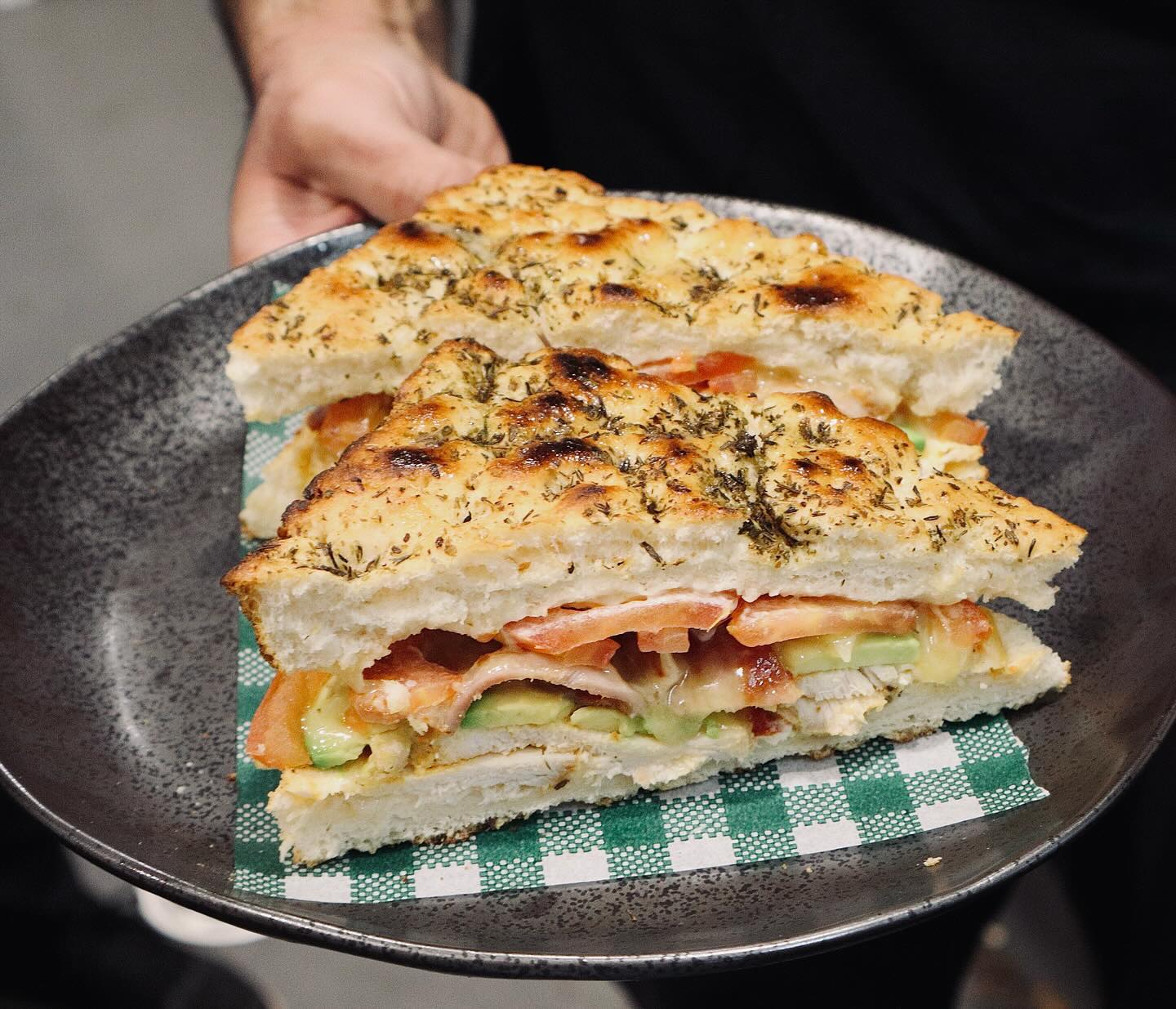 A toasted focaccia sandwich with tomato and avocado on a dark plate